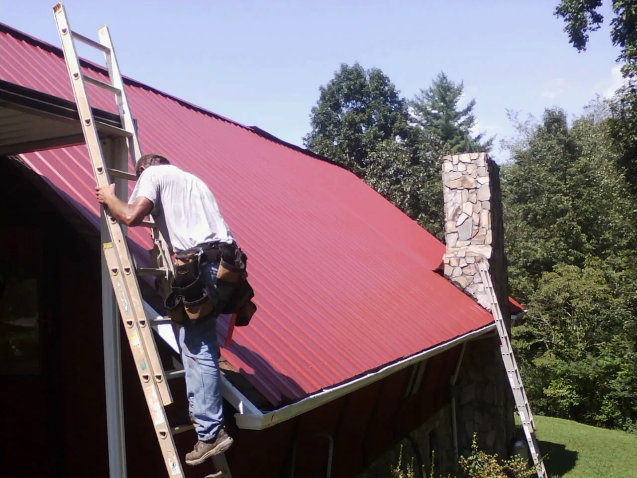 Roofer on a ladder beside a red metal roof and stone chimney — Soffit & Fascia Repair by Roofing Brothers in Ruther Glen